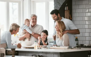 A family is gathered together in a brightly lit kitchen. A senior man holds an infant in his arms.