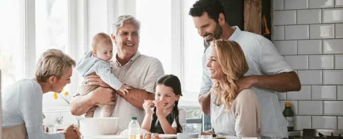 A family is gathered together in a brightly lit kitchen. A senior man holds an infant in his arms.